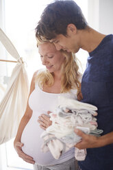 Schwangeres Paar hält Bauch und Stapel von Babykleidung im Kinderzimmer - CUF07742