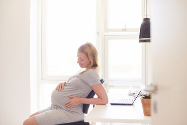 Schwangere junge Frau am Schreibtisch mit Blick auf den Bauch - CUF07771