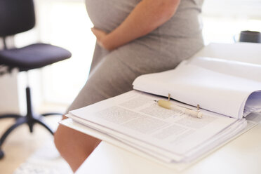 Mittelteil einer schwangeren jungen Frau, die sich an einen Schreibtisch lehnt - CUF07774