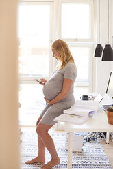 Schwangere junge Frau lehnt am Schreibtisch und schaut auf ihr Smartphone - CUF07775
