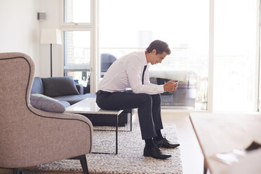 Businessman sitting on coffee table looking at smartphone - CUF07786