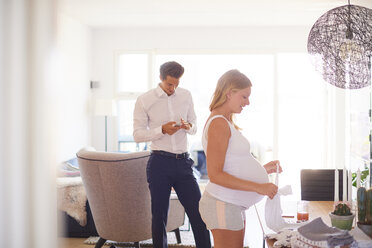 Mann schaut auf Smartphone und schwangere Freundin faltet Wäsche im Wohnzimmer - CUF07799