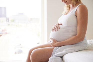 Cropped shot of pregnant young woman sitting on bed with hands on stomach - CUF07808