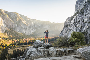 Woman on boulder looking out at valley forest through binoculars, Yosemite National Park, California, USA - CUF07880