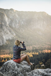 Woman sitting on boulder looking out through binoculars, Yosemite National Park, California, USA - CUF07881