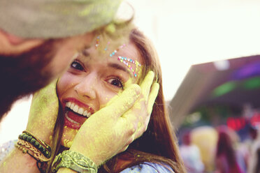 Portrait of young woman and boyfriend cupping her face with yellow chalked hand at festival - ISF01768