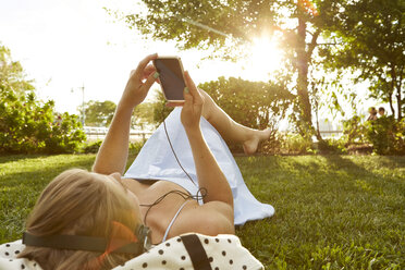 Junge Frau liegt im sonnenbeschienenen Park und wählt Musik auf ihrem Smartphone - ISF01969