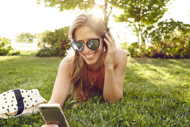 Junge Frau liegt im sonnenbeschienenen Park und hört Musik auf ihrem Smartphone - ISF01970