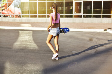 Young woman walking along road, carrying skateboard, rear view - CUF08269