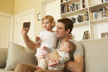 Mid adult man taking smartphone selfie with toddler and baby daughter on sofa - CUF08619