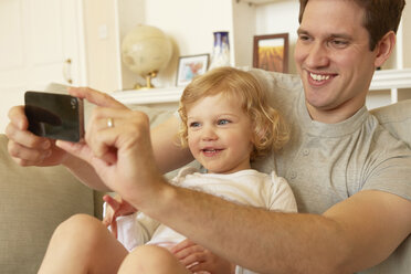 Female toddler sitting on father's knee taking smartphone selfie - CUF08620