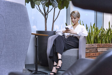 Geschäftsfrau sitzt auf einem Stuhl im Büro und benutzt ein Smartphone - CUF08751