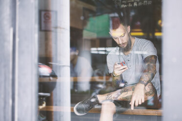 Junger männlicher Hipster auf dem Fensterplatz eines Cafés, der auf sein Smartphone schaut, Shanghai French Concession, Shanghai, China - CUF08856