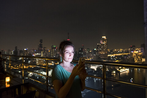 Thailand, Bangkok, Porträt einer Frau, die nachts auf einer Dachterrasse steht und ein Mobiltelefon benutzt, lizenzfreies Stockfoto