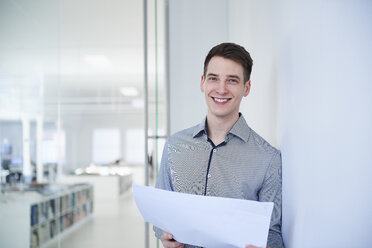 Porträt eines Architekten im Büro, der Blaupausen hält und lächelnd in die Kamera schaut - CUF09106