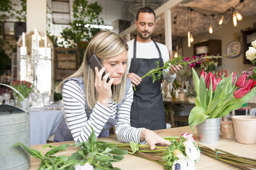 Eine Floristin telefoniert mit ihrem Smartphone, hinter ihr bereitet ein männlicher Kollege eine Blumenbestellung vor - CUF09132