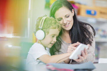 Vorschullehrerin zeigt einem Jungen mit Kopfhörern ein Mini-Tablet im Kindergarten - ZEF15467