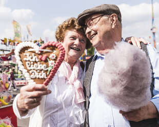 Porträt eines glücklichen älteren Paares mit Lebkuchenherz und Zuckerwatte auf dem Jahrmarkt - UUF13740