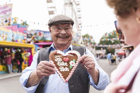 Portrait of happy senior man presenting gingerbread heart on fair - UUF13742