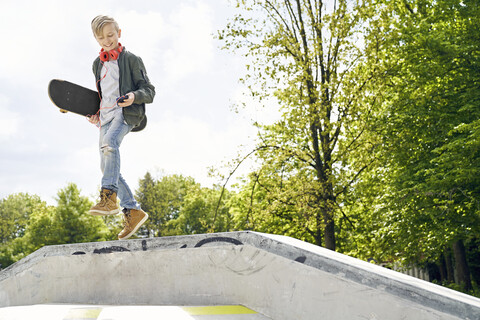 Junge mit Kopfhörern und Smartphone springt von der Rampe im Skatepark, lizenzfreies Stockfoto