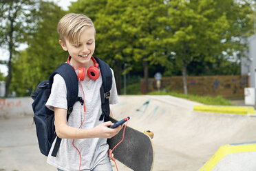 Junge mit Kopfhörern, Skateboard und Schultasche benutzt Smartphone - PDF01643