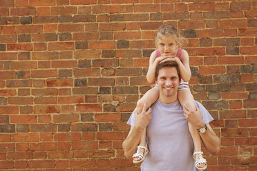 Portrait of mid adult man carrying daughter on shoulders by brick wall - CUF09280