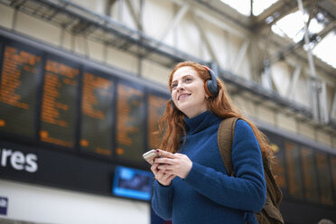 Junge Frau am Bahnhof, mit Kopfhörern und Smartphone in der Hand - CUF09289