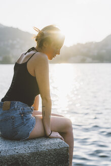 Junge Frau sitzt auf einer Mauer am Ufer und blickt auf den Comer See, Lombardei, Italien - CUF09362