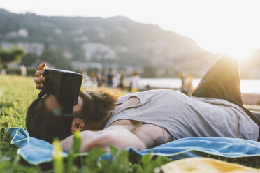 Junger männlicher Hipster, der im Gras liegt und ein Virtual-Reality-Headset betrachtet, Comer See, Lombardei, Italien - CUF09377