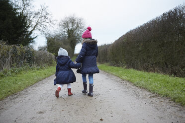 Rear view of girl and female toddler in knit hats walking along rural road - CUF09589