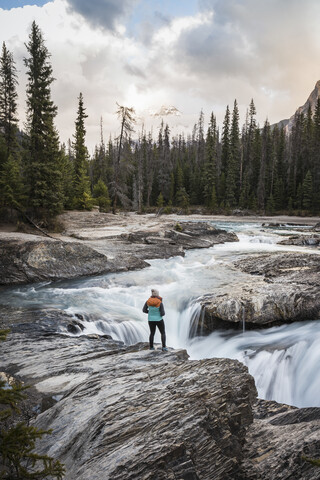 Frau steht am Rand eines Wasserfalls und betrachtet die Aussicht, Natural Bridge Falls, Kicking Horse River, Yoho National Park, Field, British Columbia, Kanada, lizenzfreies Stockfoto