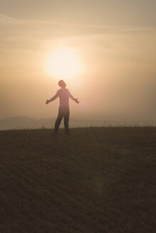 Mann steht bei Sonnenuntergang auf einem Feld, Arme geöffnet - CUF09647
