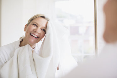 Lächelnde reife Frau trocknet ihr Gesicht mit einem Handtuch am Badezimmerspiegel - HOXF03522
