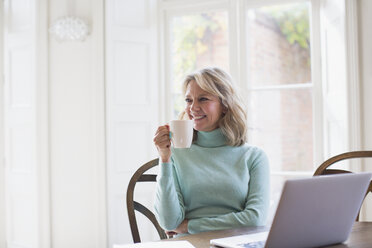 Smiling mature female freelancer drinking tea and working at laptop at home - HOXF03554