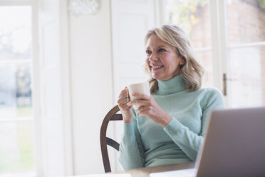 Lächelnde reife Freiberuflerin trinkt Tee und arbeitet am Laptop zu Hause - HOXF03556