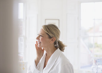 Smiling mature woman applying moisturizer to face in bathroom - HOXF03557