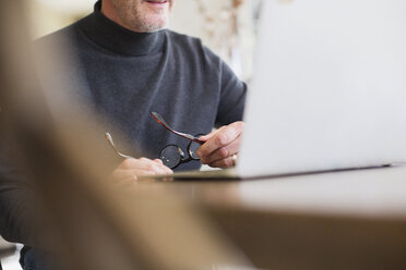 Mature male freelancer holding eyeglasses, working at laptop - HOXF03565