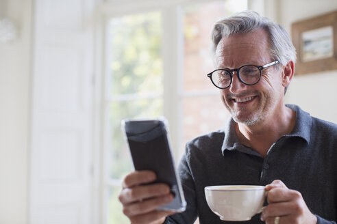 Lächelnder reifer Mann, der Tee trinkt und sein Smartphone benutzt - HOXF03603