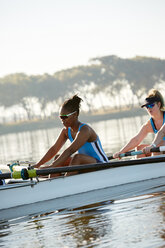 Female rowing team rowing scull on sunny lake - CAIF20653