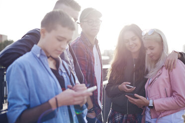 Five young adult friends looking at smartphones in sunlit city - CUF10026