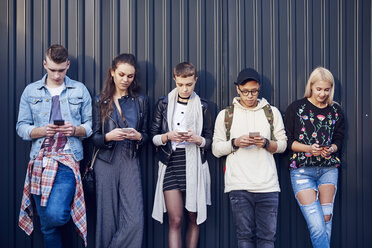 Row of five young adult friends leaning against black wall looking at smartphones - CUF10030