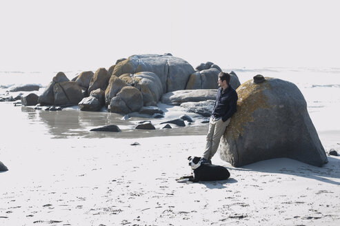 Mittlerer erwachsener Mann mit Hund, der sich an einen Felsen lehnt und auf das Meer blickt, Kapstadt, Westkap, Südafrika - CUF10188