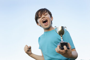 Boy making fist and holding sport trophy against blue sky - CUF10337