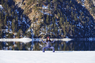 Frau in Winterkleidung übt Yoga-Pose am verschneiten Seeufer, Österreich - CUF10366