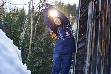Frau in Winterkleidung übt Halbmond-Yoga-Pose im Schnee bei einer Blockhütte, Österreich - CUF10372