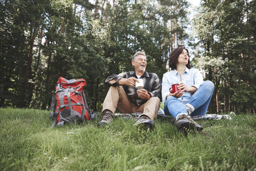 Mature couple relaxing on grass, holding tin cups, rucksack beside them - CUF10538