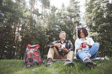 Mature couple relaxing on grass, holding tin cups, rucksack beside them - CUF10539