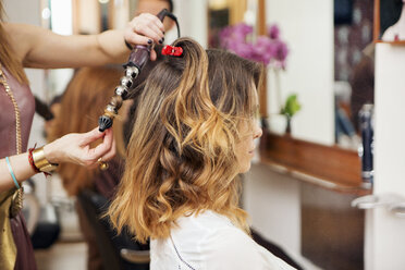 Friseurin bearbeitet das lange braune Haar einer Kundin im Salon mit einer Lockenstabzange - CUF10555