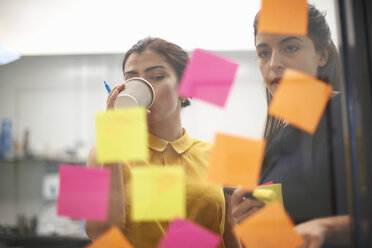 Two businesswomen drinking coffee and staring at sticky notes on office glass wall - CUF10569