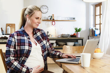 Schwangere Frau hält sich den Bauch und schaut auf einen Laptop auf dem Tisch - CUF10576